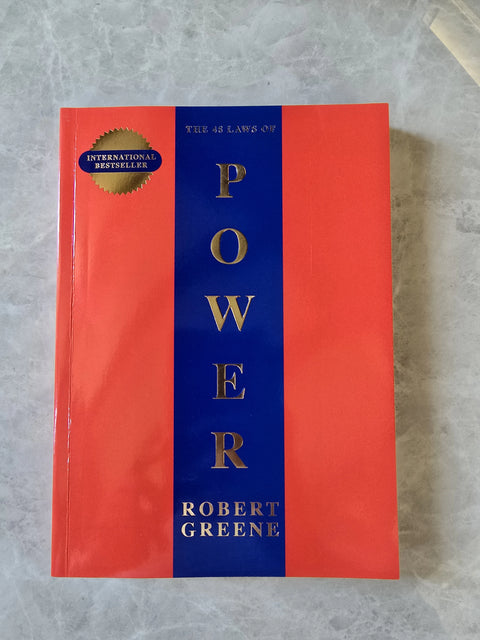 The 48 Laws of Power book by Robert Greene, featuring a red and blue cover with gold lettering, placed on a gray stone surface.